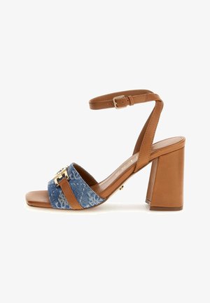 Sandal con tacón cuadrado en tono beige, con una parte superior de tela vaquera, dos correas anchas, un detalle de hebilla dorada y una correa en el tobillo ajustable.