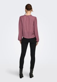 Γυναίκα με τα μαλλιά της σε κότσο, φοράει μακρυμάνικο πουκάμισο μωβ απόχρωσης, στενά μαύρα τζιν και μαύρες δερμάτινες μπότες με τακούνι, που στέκεται με την πλάτη γυρισμένη.