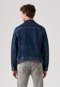 Veste en denim bleu foncé au style classique, avec des fermetures à boutons et des coutures visibles. Portée sur un jean gris clair avec des poches arrières.
