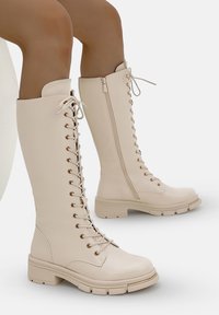 Stivali beige alti fino al ginocchio realizzati in pelle liscia. Presentano lacci, una cerniera laterale e suole spesse in gomma con un motivo di battistrada.