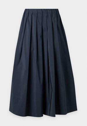 Femme debout les mains dans les poches, portant un polo blanc à manches courtes, une longue jupe plissée foncée et des ballerines noires pointues.