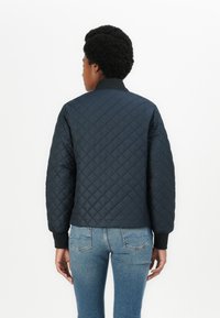 Quilted black jacket with a ribbed collar and cuffs, featuring a textured diamond pattern. Worn with light blue jeans.