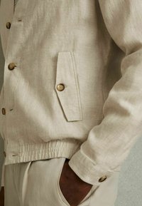 Chaqueta de lino color beige claro con botones de madera, un bolsillo en el pecho y dobladillo elástico. Textura suave con un ajuste relajado.