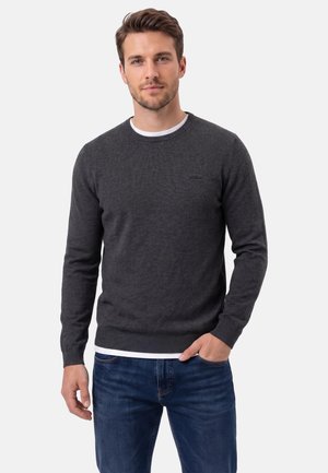 Uomo che indossa un maglione a maniche lunghe grigio scuro sopra una camicia bianca e jeans blu, in piedi con una mano in tasca davanti a uno sfondo semplice.