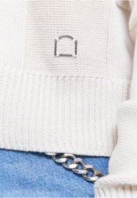 Pull tricoté crème avec détail de fermoir en métal argenté et jean bleu en denim avec une ceinture chaîne épaisse argentée.