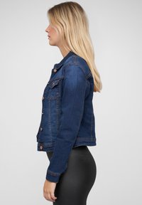 Veste en denim bleu foncé avec un design ajusté, un devant à boutons, deux poches poitrine et des accents cousus. Coupe droite à l'ourlet.