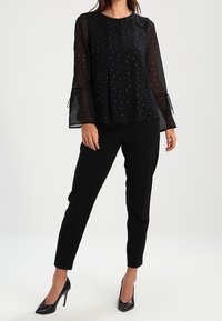 Blusa nera con maniche lunghe e trasparenti caratterizzata da un motivo a pois, abbinata a pantaloni neri sartoriali e décolleté neri a punta.
