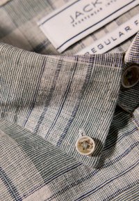 Camisa de manga corta en tejido de sarga, con un patrón a rayas en azul y beige, con un botón blanco y la etiqueta de "JACK." Corte regular.