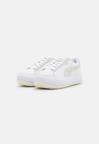 Zapatillas deportivas blancas de caña baja con suelas gruesas color beige, cordones blancos y una franja del logo de Puma en beige en el lateral, mostradas sobre un fondo liso.
