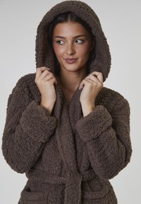 Brun huva som är tillverkad av texturerat, fluffigt material. Har stora manschetter, ett snörbälte och sidofickor. Varm och mysig design.