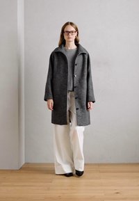 Grå uldfrakke med stor krave, knappet foran og rummelig pasform, båret over en lys grå ribbet sweater og brede hvide bukser.