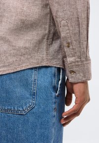 Καφέ μανσέτα μακρυμάνικου πουκαμίσου με κουμπί πάνω από την τσέπη μπλε τζιν παντελονιού, που δείχνει ένα χαλαρό χέρι και καρπό σε φωτεινό φόντο.