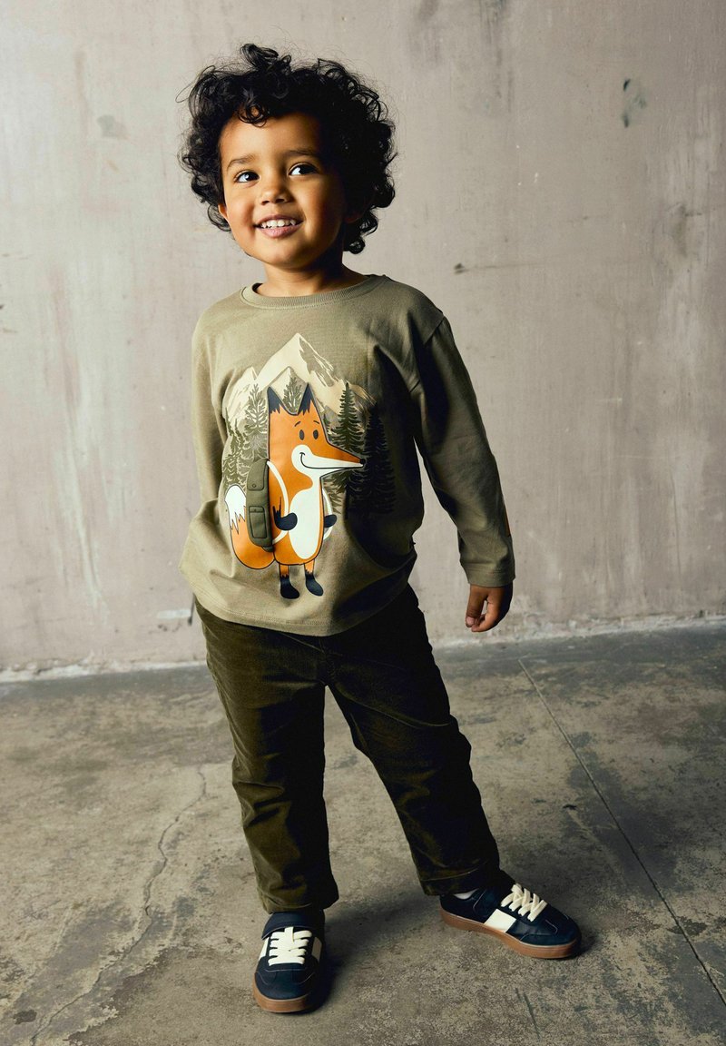 Enfant souriant aux cheveux bouclés debout sur un sol en béton, portant un sweat vert avec un imprimé renard, un pantalon foncé et des baskets bleu marine avec des lacets blancs.