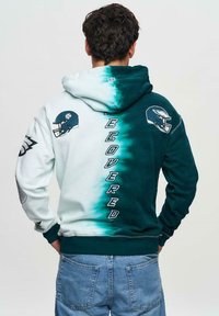 Hoodie med en gradation från vit till teal, med laglogotyper och texten "ÅTERHÄMTAD" längs ryggen i vitt. Tillverkad av bomull.