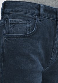 Σκούρο μπλε τζιν παντελόνι κοντινή λήψη που δείχνει την μπροστινή τσέπη, τη θηλιά της ζώνης και τις λεπτομέρειες των ραφών σε ένα άτομο που φοράει γκρι πουκάμισο.