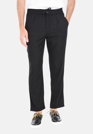 Uomo che indossa pantaloni neri su misura con vita regolabile a coulisse e scarpe di pelle nere con lacci gialli, in piedi con le mani nelle tasche.