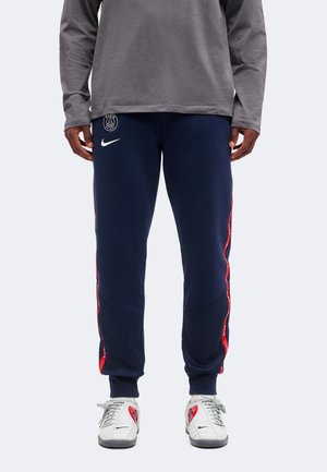 Άτομο που φοράει ναυτικό μπλε φόρμα PSG της Nike με κόκκινες ρίγες στο πλάι, γκρι μπλούζα με μακριά μανίκια και λευκά αθλητικά παπούτσια Nike με κόκκινες λεπτομέρειες.