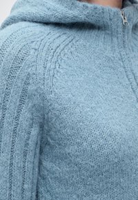 Pull en tricot bleu clair doux avec des motifs texturés et une fermeture éclair argentée visible près du cou.