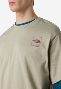 Krémové tričko s krátkým rukávem, na levé části hrudi má bordó čtvercové logo. Pod tím je částečně viditelná modrá vrstva s dlouhým rukávem.