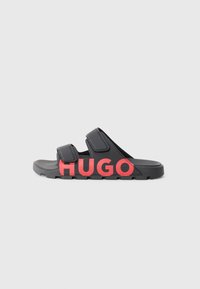 Sandale negre cu două curele reglabile, având un logo roșu îndrăzneț "HUGO". Talpă texturată pentru aderență, material ușor. Vedere din lateral.