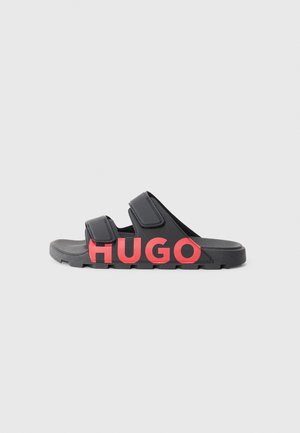 Schwarze Sandalen mit zwei verstellbaren Riemen und einem auffälligen roten "HUGO"-Logo. Strukturiertes Profil für besseren Grip, leichtes Material. Seitenansicht.