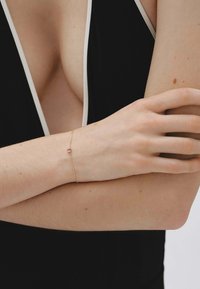 Primer plano de una persona que lleva una prenda negra de escote en V profundo y una delicada pulsera de oro con una pequeña perla roja y blanca en la muñeca.