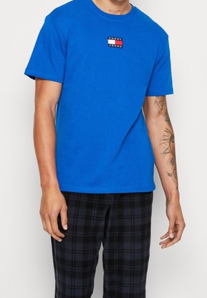 Mann trägt ein leuchtend blaues Tommy Jeans T-Shirt mit kleinem Logo auf der Brust und schwarze karierte Hosen, zeigt Tattoos am linken Arm.