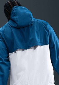 Personne aux cheveux tressés porte une veste à capuche bleue et blanche, vue de dos sur un fond gris foncé.