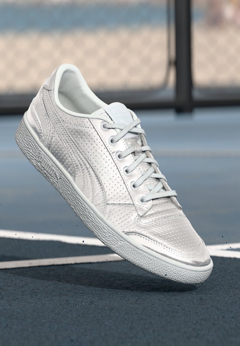 Hvid Puma-sneaker med snørebånd, perforeret tekstur og gummisål, der svæver over en blå sportsbane med hvide linjer.