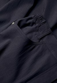 Donker marineblauwe stof met een gladde textuur, voorzien van een elastische tailleband en een zijzak met een metalen drukknopdetail. Eenvoudig, functioneel ontwerp.