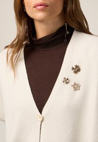 Witte cardigan met een v-hals en knoopsluiting, gedragen over een donkerbruine coltrui. Drie decoratieve broches: ster- en bloemvormen.