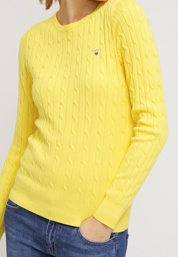 Pull jaune en maille torsadée avec manches longues et petit logo sur la poitrine, porté avec un jean bleu, main dans la poche.