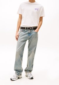 Biała koszulka z fioletowym logo, jasnoniebieskie dżinsy, czarny pasek z srebrnymi elementami oraz szare sneakersy z zielonymi akcentami.