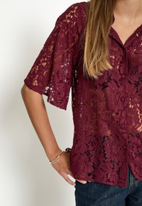 Blusa in pizzo bordeaux con maniche corte, motivo floreale, chiusura con bottoni e lacci all'orlo, realizzata in un tessuto leggero e testurizzato.