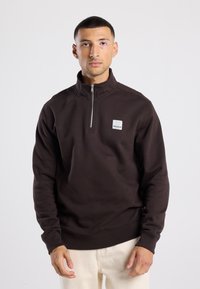 Brown zip-up sweatshirt with high collar, featuring a patch with "BROOKLYN" text, made from a smooth fabric. Worn over light-colored pants.