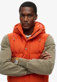 Gepolsterte Weste in leuchtendem Orange mit Kapuze, ausgestattet mit Druckknöpfen, Kordelzug-Details und einem kontrastierenden beigefarbenen Pullover darunter.