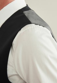 Schwarze Weste aus glattem Stoff mit einem durchsichtigen grauen Panel an der Schulter, kombiniert mit einem weißen Hemd. Klare Linien und maßgeschneiderte Struktur.