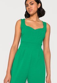 Macacão verde com decote quadrado, corpete estruturado e alças largas. Feito de tecido suave com costuras verticais e pernas ligeiramente largas.