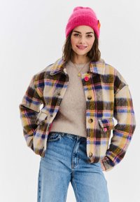 Kostkovaná bunda z vlněné směsi v žluté, modré, hnědé a růžové barvě, s knoflíky a límcem. Nositá přes béžový svetr a džíny.