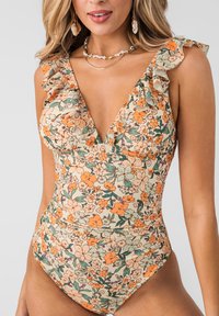 Vrouw draagt een bloemenbadpak met ruches aan de schouderbandjes en gelaagde kettingen, tegen een eenvoudige lichtgrijze achtergrond.