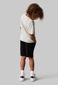T-shirt a righe a maniche corte color crema con righe blu scuro, abbinato a pantaloni corti neri. Calzini bianchi e scarpe da ginnastica nere completano l'outfit.