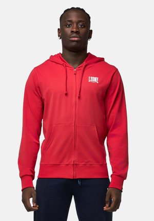 Uomo di colore che indossa una felpa con cappuccio rossa con zip e il logo "LEONE 1947" sul petto e pantaloni scuri, in piedi contro uno sfondo semplice.