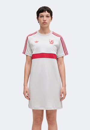 Persona de pie, de frente, con un vestido gris de manga corta con rayas rojas en los hombros y una banda roja en el pecho.