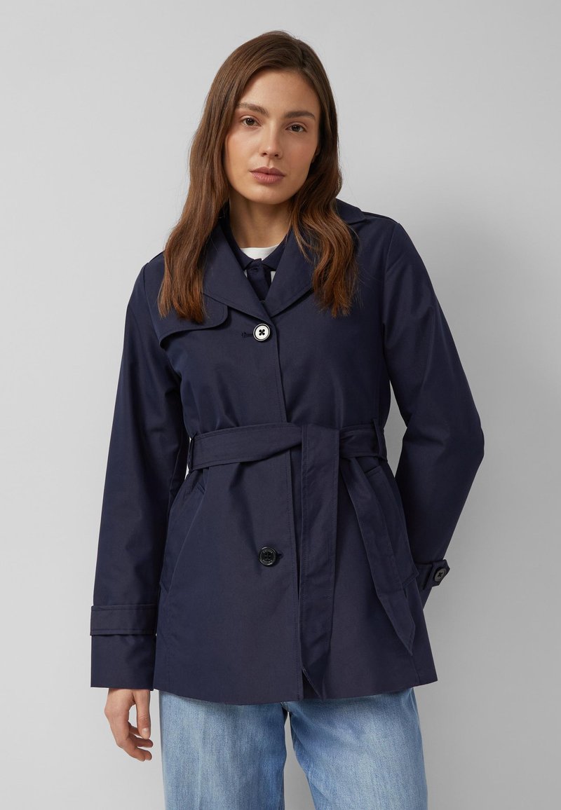 Frau mit braunen Haaren, die einen marineblauen Trenchcoat mit Gürtel über einem weißen Oberteil und hellblauen Jeans trägt, vor einem einfachen grauen Hintergrund stehend.