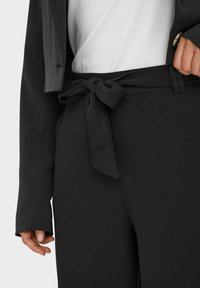 Pantalon noir en tissu texturé, avec une ceinture nouée et une silhouette ajustée, assorti à un haut blanc et un blazer foncé.