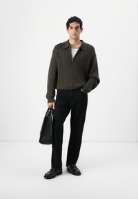 Ανδρας φορώντας καφέ φούτερ με κλείσιμο, μαύρο παντελόνι, μαύρα παπούτσια, κρατώντας μια μαύρη δερμάτινη τσάντα, στέκεται μπροστά σε λευκό φόντο.