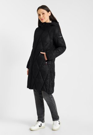 Zwarte gewatteerde parka met een capuchon, voorzien van een diagonaal patroon, twee zijzakken en een ritssluiting, gedragen over grijze jeans en witte sneakers.