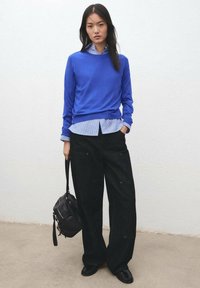 Maglione blu a maglia indossato sopra una camicia a righe azzurre, abbinato a pantaloni neri larghi. Tiene una piccola borsa a tracolla nera.