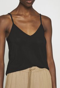 Débardeur noir à encolure en V et bretelles fines, en tissu léger, associé à un pantalon beige à cordon de serrage, avec une coupe décontractée.