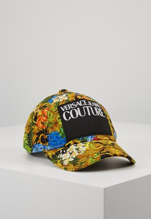 Casquette avec imprimé floral coloré et baroque, patch rectangulaire noir affichant "Versace Jeans Couture" en lettres blanches, posée sur une surface blanche.